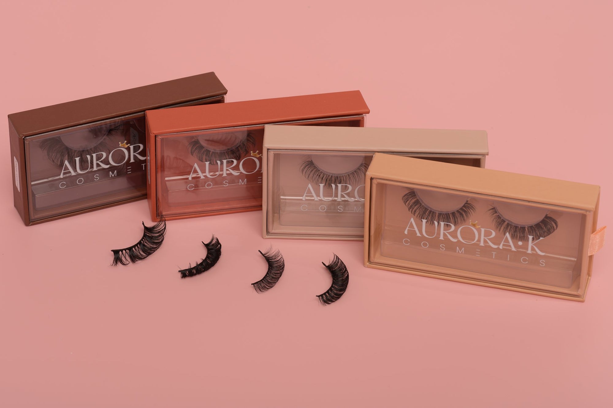 A group of four lash boxes in warm brown colors with the lashes (classic, volume, volume and mega volume) laid out by the side from Aurora K Cosmetics